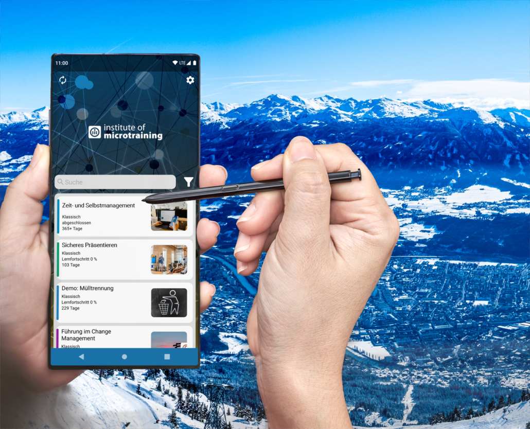 2 Hände halten ein mobiles Device, worauf die Trainingsübersicht auf dem Startscreen der Microtraining®-App angezeigt ist, im Hintergrund ein Panoramablick auf Innsbruck.